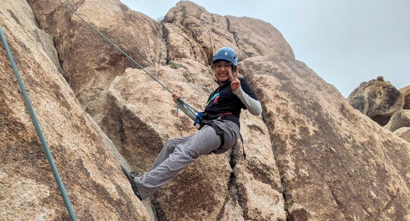 A person wearing safety gear is secured by ropes as they pause climbing a rock wall to smile and give a peace sign for the photo.